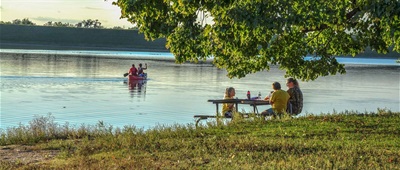 Two people paddle a canoe on the lake, while in the foreground, two youth and an adult enjoy a picnic at a park’s picnic table under a leafy shade tree.