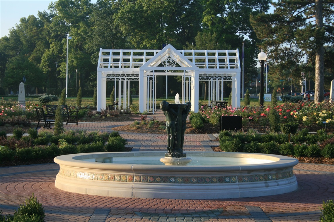 The Joy Fountain marks the center of the rose garden, framing it in the background is a  Rose inspired Art Deco Gazebo. Roses in yellow, peaches, and pinks fill in the beds behind the fountain.