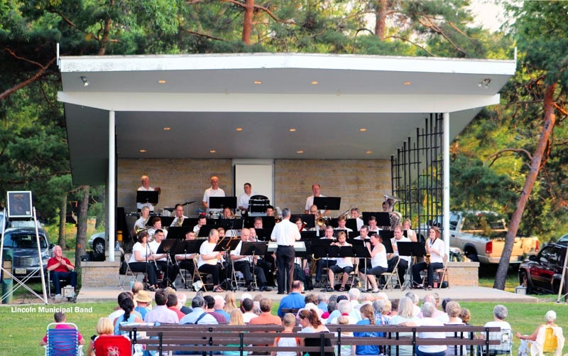 The Lincoln Municipal Band perform in the Shildneck Band Bandshell on a sunny afternoon against the backdrop of established conifers. 