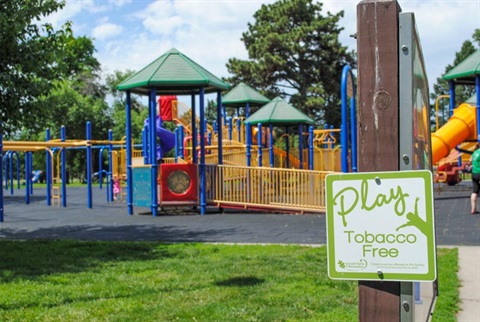 A sign reads, “Play Tobacco Free” in the foreground. Green roofs of various heights mark shaded play platforms. Yellow rails show a maze of ways to explore and engage with the variety of colorful playground equipment.