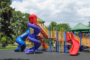 Part of Antelope park’s large playground, a tall tower leads to a blue slide, and a purple spiral slide. There is a lower, smaller red slide. The play surface is rubber tiles and large trees provide shade to the nearby picnic tables and benches.