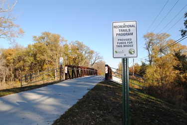 Park’s sign reads “Recreational Trails Program Provided Funds For This Bridge”, behind the sign, is a new hiker biker bridge.