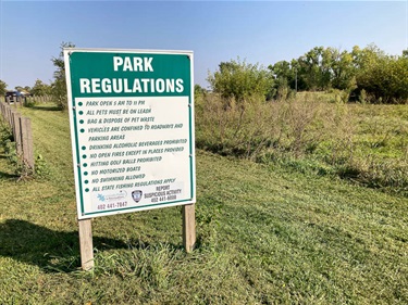 Park sign with relevant park regulations listed. Lincoln Parks and Recreation contact: 402-441-7847. Behind the sign are both mowed and un-mowed park greenspace.