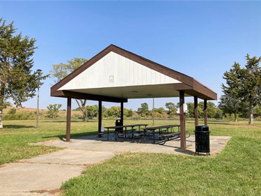The open shelter at Bowling Lake is located on a concrete slab. It has two long picnic tables as well as garbage cans. The shelter is located near several large evergreens.
