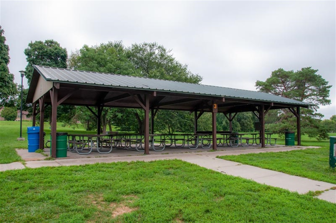 Holmes Lake Shelter number 3 is located on a concrete slab connected to the park’s paved trail. There are picnic tables for a large group, open space, and large trees nearby.