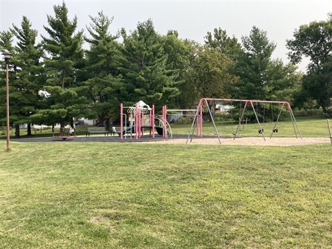 Play area with multi level structure, two slides and a variety of climbing elements, on a rubber tiled surface. On the sand area are belt swings and bucket swings. Tall shade trees, benches, and an open grass area surround the playground.