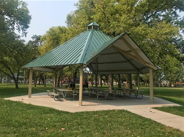A large open shelter with a vaulted green roof sits on a concrete slab connected to the paved walking trail nestled among established trees.