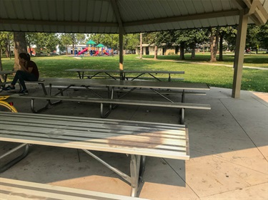 Underneath the open shelter are metal picnic tables on a concrete surface. The playground and walking trail are in the background.