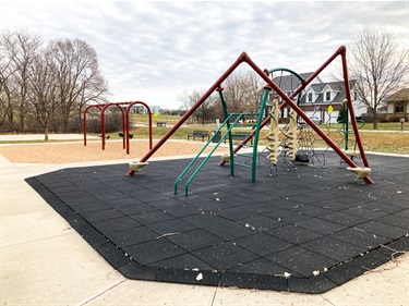This play structure features a variety of unique climbing elements on a rubber tiled area, adjacently there is a swing set with both bucket and belt swings. The areas have paved paths around them, and benches nearby.