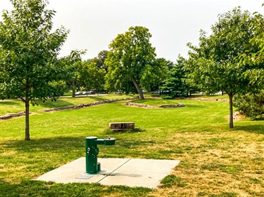 A park’s drinking fountain is framed by the varying sized trees, and a large open green space.