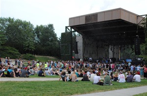 A crowd sits in the green space in front of the Pinewood Bowl stage. Large pine trees fill in the background.