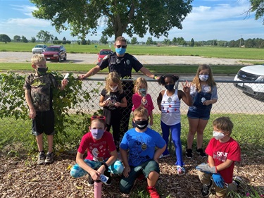 A group of campers pose with a visiting police officer outside. The campers are all either wearing or holding their silver badge stickers. All are wearing face coverings.