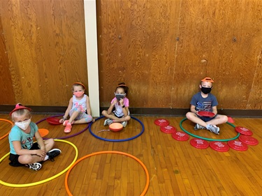 A group of four campers play an indoor, socially distant, game with hula hoops and frisbees. The campers are sitting in their own hula hoops. All campers have a cone on their head and a cloth face covering.