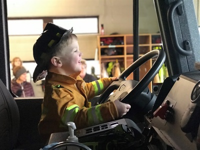 Child dressed in firefighter clothes pretending to drive a fire engine.
