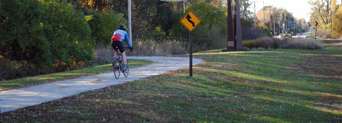 A single biker rides away from the viewer on a paved trail. In front of them the trail turns into a wooded area.
