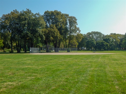 Large shade trees stand behind the infield, the green outfield fills in the foreground.