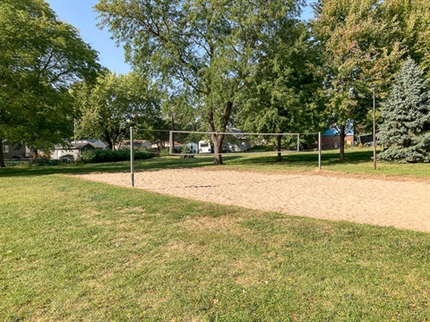 A sand volleyball court located near a group of trees providing partial shade.