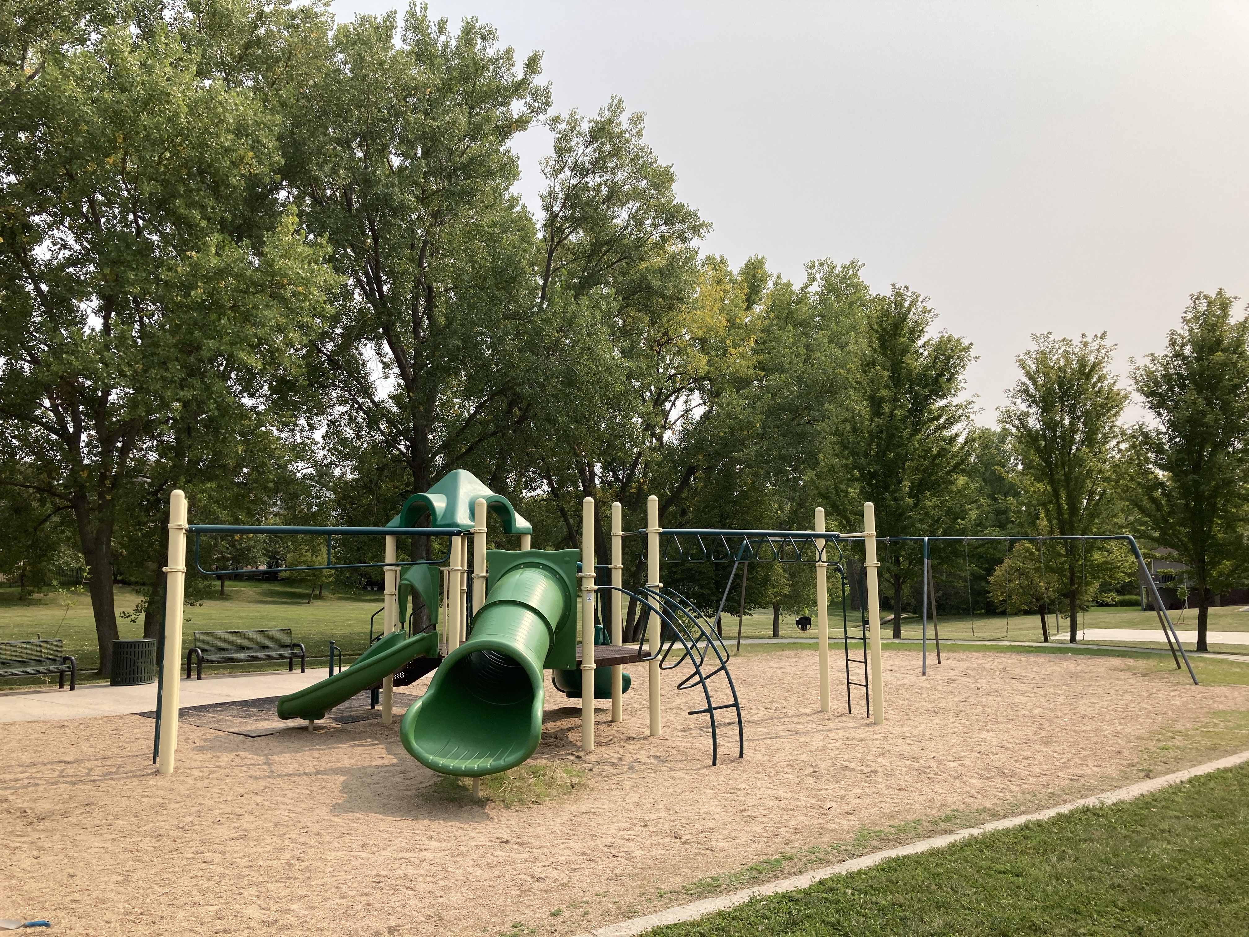 A playground area on sand featuring a multilevel structure with slides and varying climbing elements. There is a separate structure with belt and bucket swings. Benches are located nearby under shade trees. 