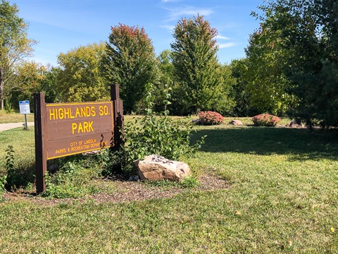 Brown park sign reads, “Highlands So. Park, City of Lincoln, Parks & Recreation Department” behind the sign are hardy perennials, a tree line, and a park’s trail.