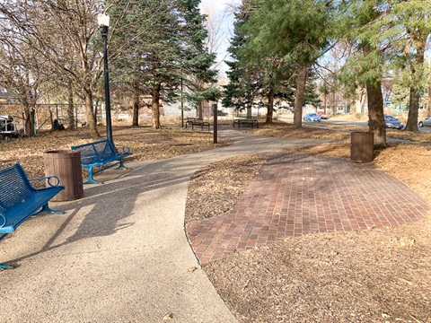 A paved path weaves through established evergreens and park benches in this pocket park.