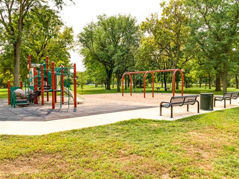 This play area is nestled among large established shade trees. The structure is multi level with a variety of climbing features and slides on rubber tiles. Adjacent, is a sand covered area with belt and bucket swings. 