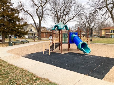 A small, multi level, playground with a slide and several different climbing elements. There is a path around the playground through large shade trees and benches.