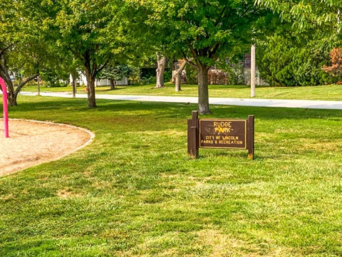 A brown park’s sign reads, “Rudge Park, City of Lincoln Parks and Recreation, behind it a row of trees frames the road.