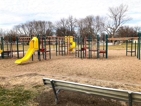 Sunrise park playground is a larger multi level structure with climbing elements and slides. The play area has a sand surface, and benches are located nearby. 