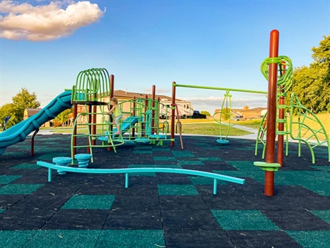 A young child plays on brightly colored climbing structures and slides on a black and teal rubber play surface in a neighborhood park.