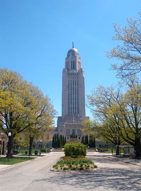Large shade trees with spring foliage frame the Nebraska Capitol building against a clear blue sky.