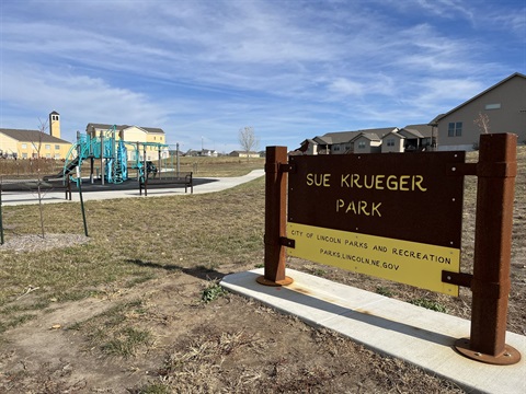 A photo of the metal signage for Sue Krueger Park with a playground and grass in the background.