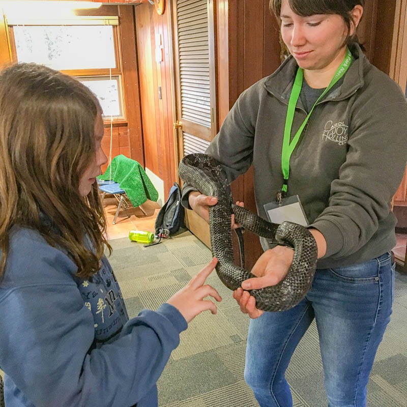 Nature Center Staff handles a snake for a student to engage with. The student gingerly places one finger on the snake.