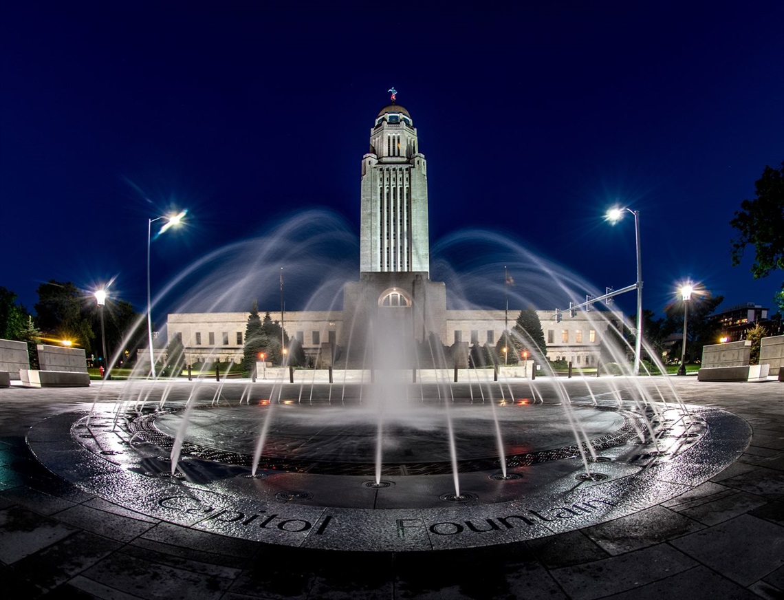 A night-time image of the the Capitol Building at night with lights reflecting the water shooting from the state seal fountain  