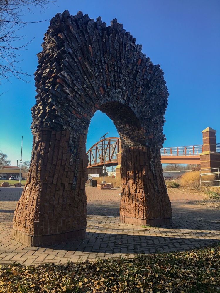 An archway made of varied sized and colored bricks, in front of a bridge