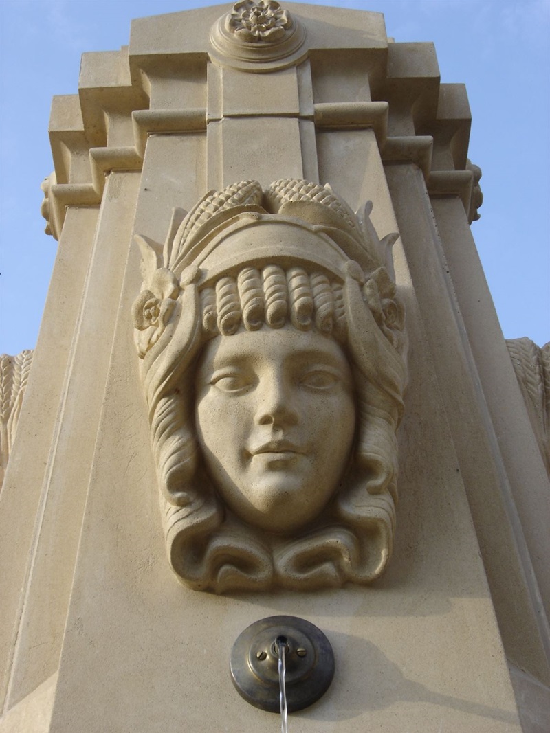 the head of a young woman wearing a corn headdress on the column of a fountain