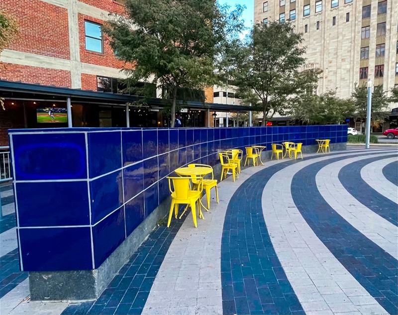 A long wall, covered in blue glazed tiles, with yellow chairs and tables in front of it. 