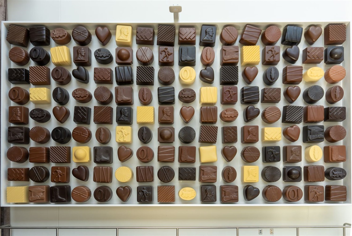 A larger than life sculpture of a box of chocolates arranged on a wall in a grid of 16 by 9 pieces of chocolate. A few pieces have designs of Abraham Lincoln, the sower, corn, a train, a basketball, a cowboy boot and a grad cap and diploma. 