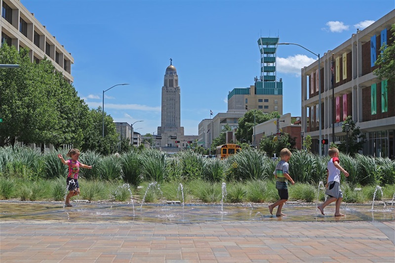 Children playing in the Imagination Fountain with part of a downtown Lincoln skyline in the background.
