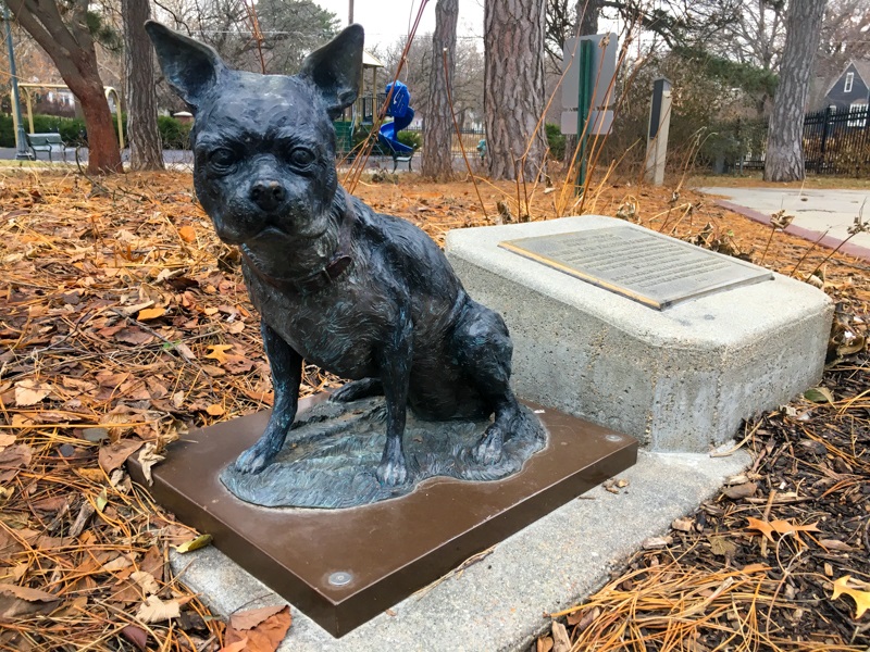 a small metal dog on a metal pedestal in front of a plaque, playground and fallen leaves