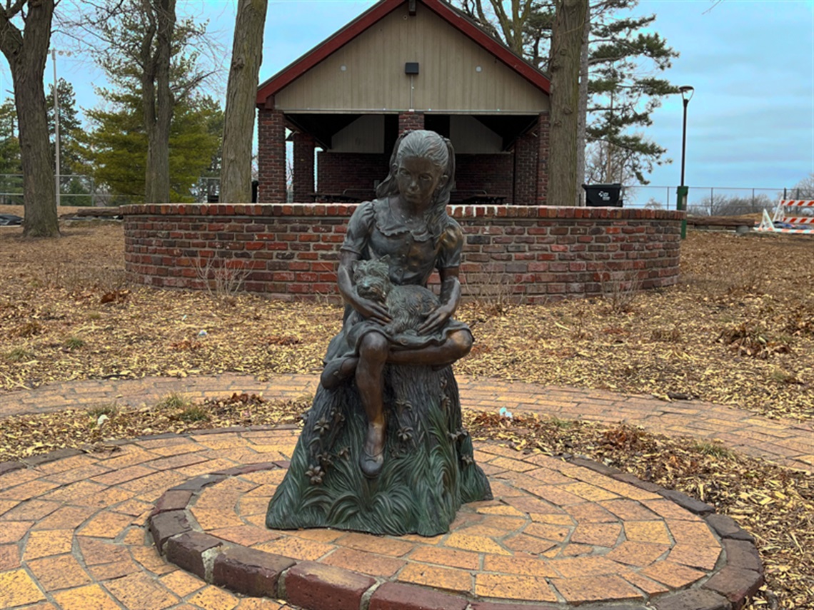A statue of a young girl with a dog in her lap, sitting on a metal stump in front of a small shelter. 