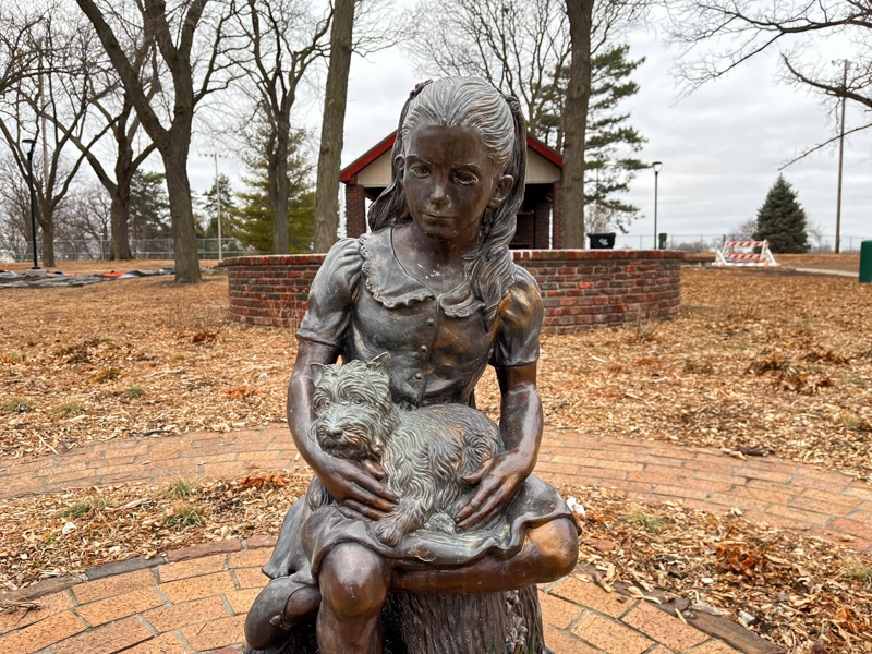A statue of a young girl with a dog in her lap, sitting on a metal stump in front of a small shelter. 