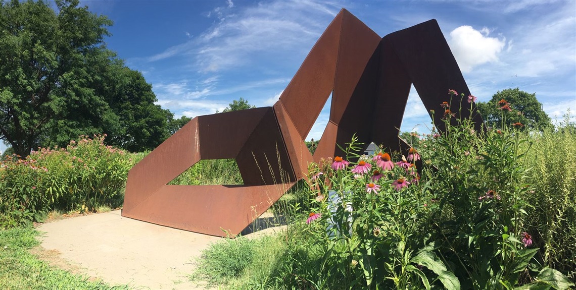 Pink flowers and grasses that block the view of part of the large geometric steel sculpture