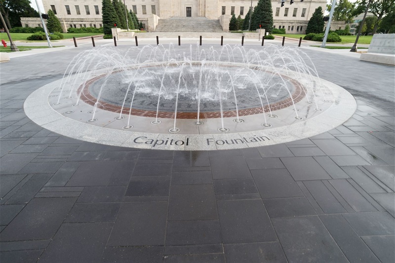 A round fountain with streams of water coming out of the pavement with the words Capitol Fountain engraved on the outer ring.
