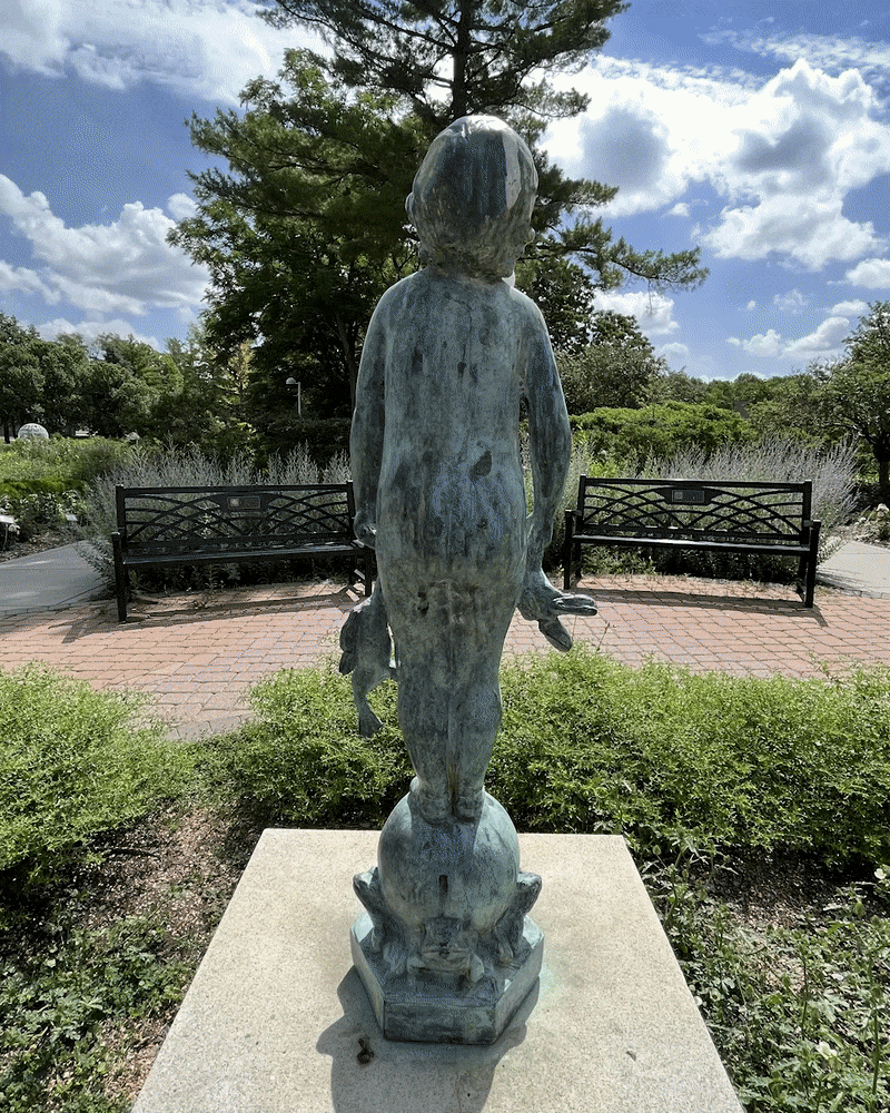 Rotating around a sculpture of a small child holding a frog in each hand with a smile on his face looking up towards the sun. 