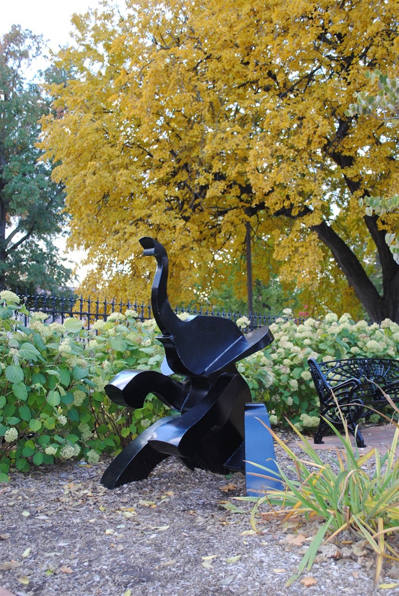 A small metal elephant amid the flowers and a yellowing tress in fall