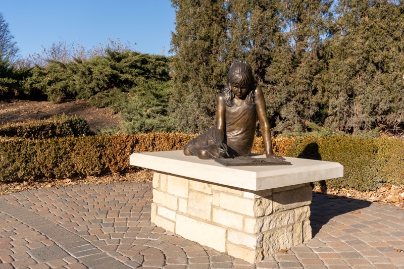 On a limestone base sits a bronze figure of a young girl looking at a book, surrounded by flora. 