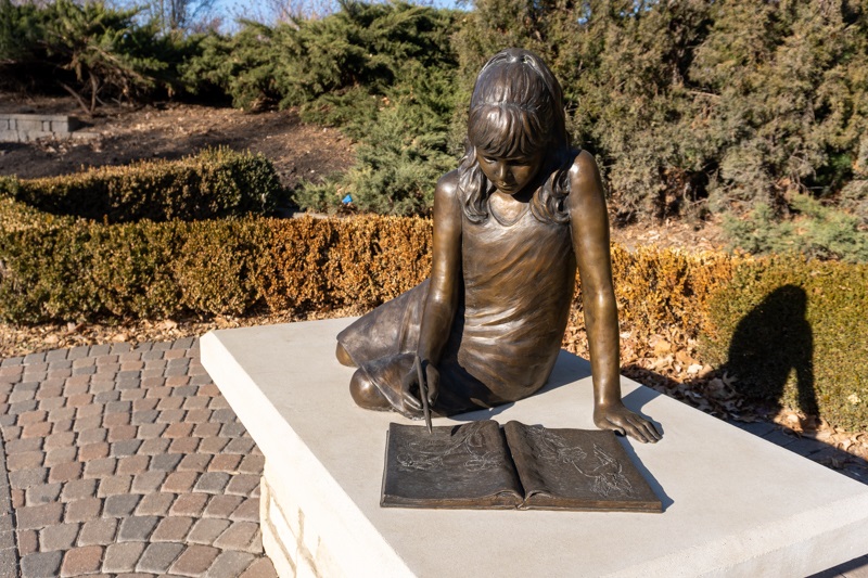 A bronze figure of a young girl drawing in her sketchbook, with bushes, and trees in the background.