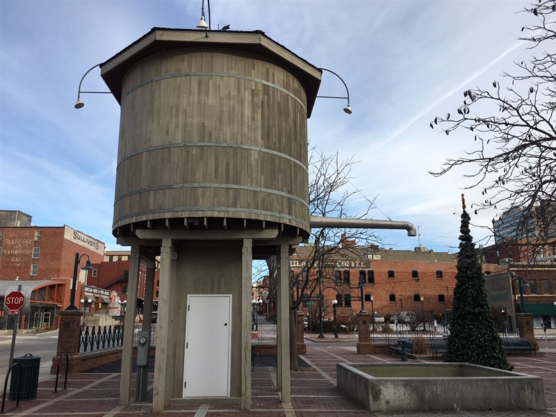 The backside of the watchtower fountain at Iron Horse park, shows the barrel like wooden water container and the pipe sticking out of the side. 