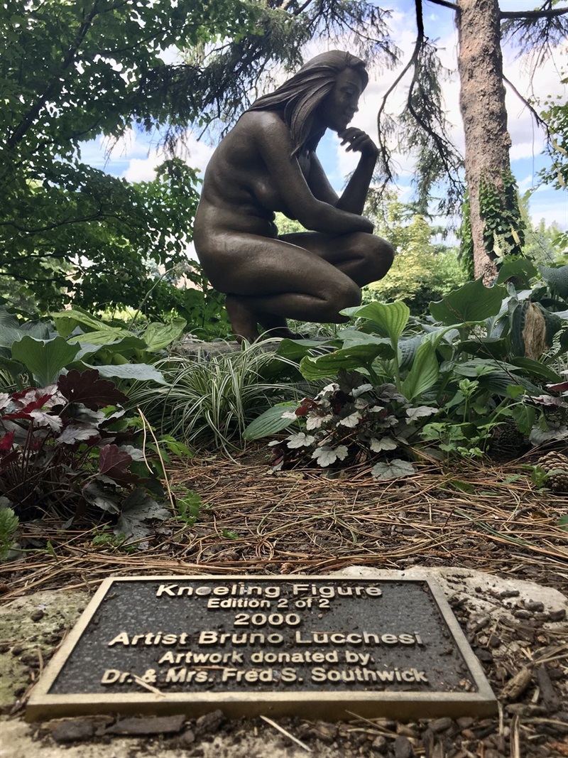 A metal figure of a woman kneeling, looking towards the ground in contemplation. The plaque for the sculpture is in the foreground. 