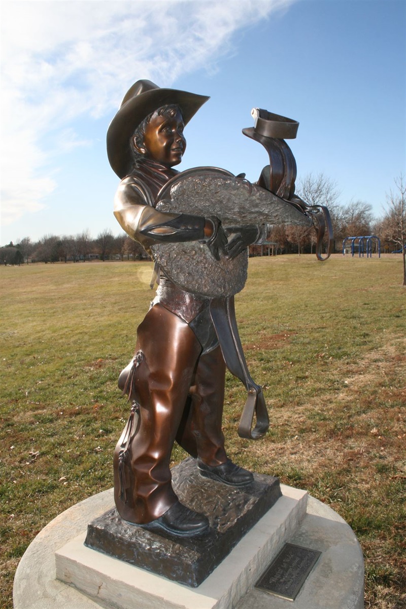 A bronze statue of a young cowboy holding his saddle with excitement on his face. A field of grass and a park is in the background. 